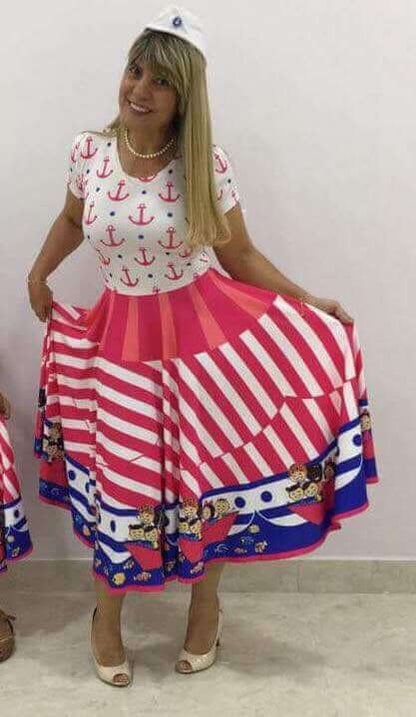 Modelo em fundo cinza veste vestido com âncoras e bolinhas azuis na blusa, saia listrada e barco com crianças na barra.
